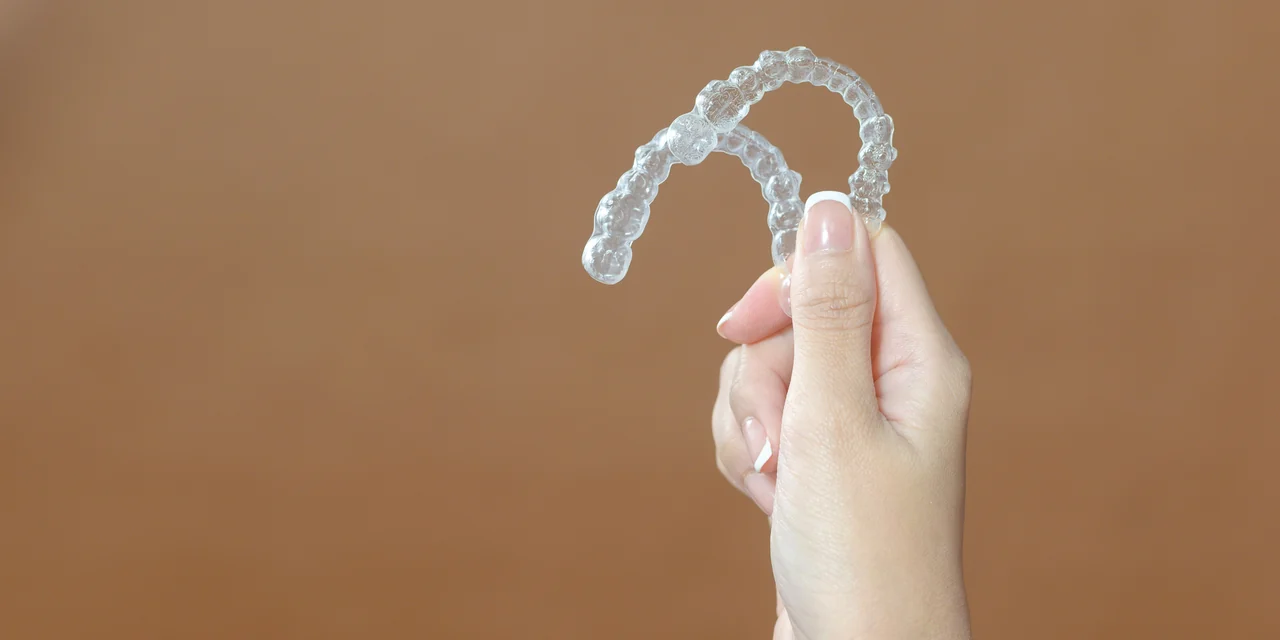 A clear orthodontic aligner held in one hand against a warm brown background